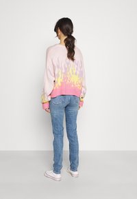 Rosa stickad tröja med flamamönster i lila och gult vid fållen, kombinerad med ljusblå jeans och vita sneakers.