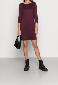Robe bordeaux longueur genou avec manches trois-quarts en tissu lisse. Portée avec des bottines noires et un petit sac à main noir.