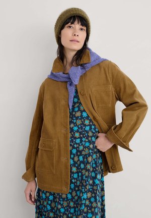 Brun kordfløjljakke med store frontlommer, båret over en mørk blomstret kjole, suppleret med et lilla tørklæde og en grøn strikket hat.