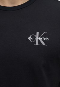 Μαύρο T-shirt με μικρό λευκό και γκρι λογότυπο Calvin Klein στο επάνω αριστερό μέρος του στήθους, δείχνοντας κοντινή λήψη του λαιμού και του άνω κορμού.