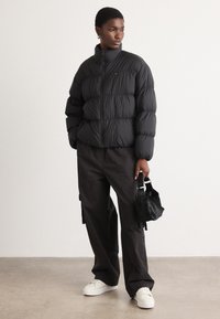 Svart pufferjakke med høy krage og glidelås foran, kombinert med svarte cargobukser og hvite joggesko. En liten svart håndveske blir holdt.