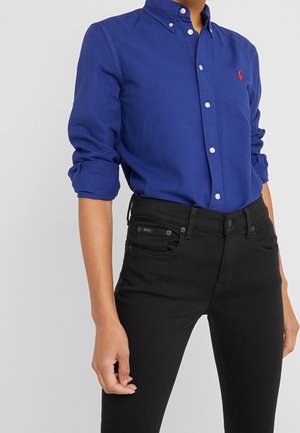 Personne portant une chemise à manches longues bleu royal boutonnée avec les manches retroussées et un jean noir ajusté avec un logo Polo sur la poche.