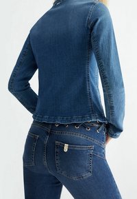 Osoba v přiléhavé tmavě modré džínové bundě a džínách s šněrováním v pase, zobrazená ze zad.