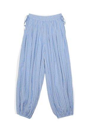 Donna con capelli ricci che indossa un top bianco senza spalline, pantaloni larghi a righe blu e bianche e tacchi bianchi aperti, in piedi contro una parete chiara e semplice.