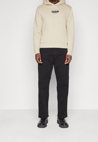 Felpa beige con logo nero, abbinata a pantaloni neri a gamba dritta e scarpe nere con suole testurizzate. Elementi di design semplici e moderni.