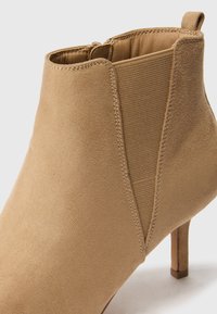 Beige nilkkuri, joka on valmistettu sileästä mokasta ja siinä on elastiset paneelit. Kenkässä on kapea korko ja takana vetoketju, joka helpottaa pukemista.