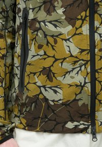 Τζάκετ με καμουφλάζ σε αποχρώσεις πράσινου, κίτρινου και καφέ· διαθέτει μαύρη τσέπη με φερμουάρ και υφασμάτινη υφή.