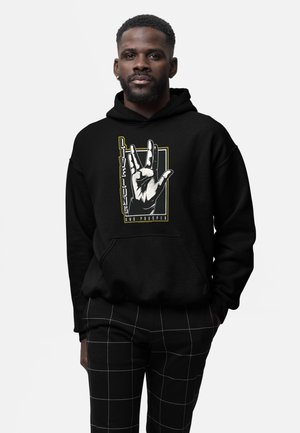 Mężczyzna w czarnej bluzie z kapturem z grafiką dłoni „Live Long and Prosper”, w połączeniu z czarnymi spodniami w białą kratę.