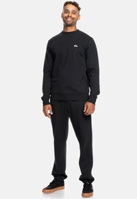 Czarny sweatshirt z okrągłym dekoltem z małym białym logo na piersi, w zestawie z czarnymi dresami i czarnymi trampkami z brązowymi podeszwami.