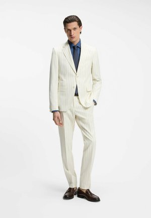 Mann trägt einen cremefarbenen Nadelstreifenanzug mit blauem Hemd, navy gepunkteter Krawatte und dunkelbraunen Loafers, steht mit einer Hand in der Tasche.