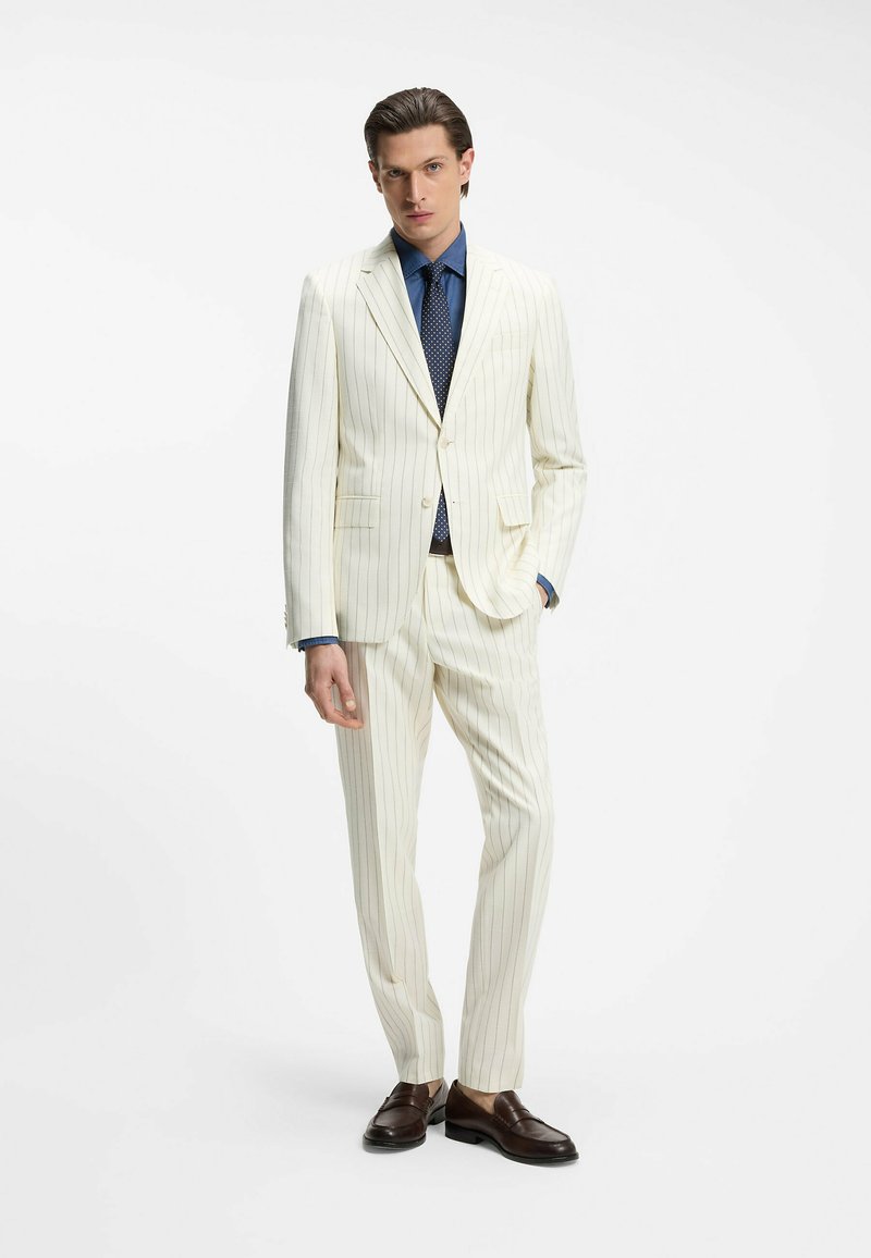 Homme portant un costume crème à fines rayures, une chemise bleue, une cravate bleu marine à pois et des mocassins marron foncé, debout avec une main dans la poche.