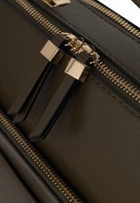 Oliivinvihreä laukku, jossa on kultainen vetoketju ja mustat nahkavetimet. Teksturoitu materiaali näkyvissä; tyylikäs, jämäkkä muotoilu.