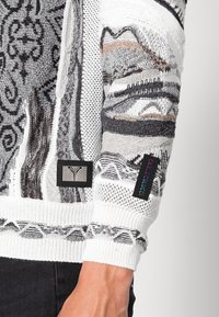 Pull en tricot avec un mélange de motifs noirs, blancs et gris, des manches texturées, et des patchs logo sur le bras et l'ourlet.