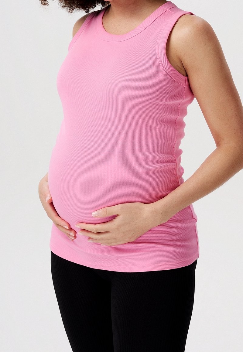 Kobieta w ciąży ubrana w różowy top bez rękawów i czarne spodnie, delikatnie obejmująca brzuch na jednolitym jasnym tle.