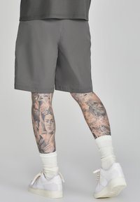 Benen met gedetailleerde zwarte en grijze tatoeages die gezichten en muzieknoten tonen, gekleed in grijze shorts, witte sokken en witte Nike sneakers op een grijze achtergrond.