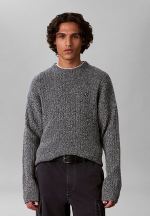 Jeune homme aux cheveux bouclés mi-longs portant un pull en laine gris et un pantalon foncé sur un fond gris uni.