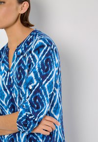 Blau-weiß gemustertes Hemd mit tiefem V-Ausschnitt, 3/4-Ärmeln und Knopfdetail an den Ärmeln, aus leichtem Stoff gefertigt.