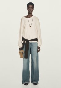 Heller beige, oversized Pullover mit einem braunen Gürtel, kombiniert mit weit geschnittenen, hellblauen Jeans. Schwarze Sandalen und ein braunes Accessoire in der Hand.
