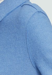 Jasnoniebieska tkanina z bliska, z teksturową powierzchnią, ukazująca lekko luźny szew na ramieniu i swobodny krój odzieży.