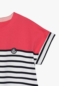 Koszulka w paski z koralowym górnym fragmentem, czarno-białymi poziomymi paskami na dole oraz okrągłym logo z żaglówką na lewej piersi.