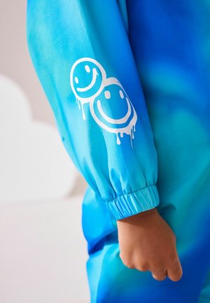 Blaues Gradientenjackett mit elastischen Bündchen, das zwei weiße, tropfende Smiley-Gesicht-Grafiken auf dem Ärmel aufweist. Glattes, leichtes Material.