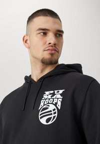 Sudadera negra con cordón, que presenta un estampado gráfico en blanco que incluye "PUMA HOOPS" y un logotipo de baloncesto. Tejido texturizado, ajuste informal.