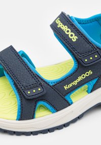 Mørkeblå sandaler med gule detaljer, justerbare stropper og teksturert såle. Har pustevennlig mesh-fôr og merkevarelogo. Lett materiale.