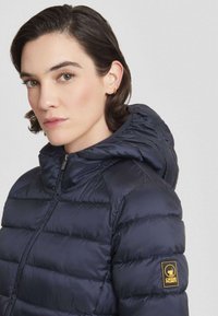 Jong persoon met kort donker haar, gekleed in een donkergroene gewatteerde jas met capuchon en een logo-patch op de mouw, kijkend iets naar links.