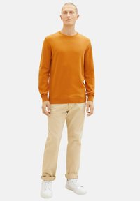 Suéter de punto naranja, cuello redondo, mangas largas; combinado con chinos beige y zapatillas blancas; textura suave, estilo ajustado.