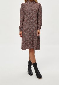 Femme portant une robe florale à manches longues, col, longueur genoux, avec des bottes à talons noires mi-mollet sur un fond blanc.