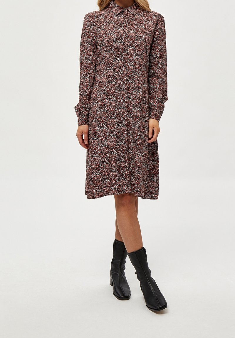 Femme portant une robe florale à manches longues, col, longueur genoux, avec des bottes à talons noires mi-mollet sur un fond blanc.