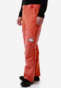Pantaloni impermeabili arancioni con finitura testurizzata, dotati di tasche laterali e di un logo bianco prominente sulla coscia. Abbinati a calzature nere.