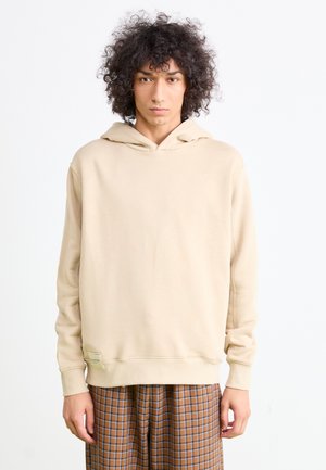 Young person with curly hair wearing a beige hoodie and brown plaid pants, standing against a plain white background.