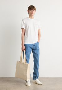 Beige canvas tote bag i rektangulær form med to håndtak og frontlomme, parret med en hvit t-skjorte og blå jeans.