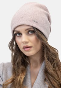 Gorro de punto rosa claro con un patrón texturizado y un pequeño acento de logo metálico. Diseño suave y holgado, adecuado para el uso casual.