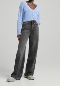 Světle modrý chlupatý kardigan s výstřihem do V a šesti knoflíky, doplněný o černé džíny s vysokým pasem a robustními bežovými teniskami.