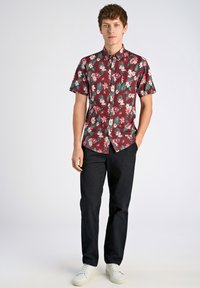 Korte mouwen, bordeauxrood bloesjeshemd met groene bladeren, gecombineerd met zwarte broek en witte sneakers. Voorpand sluiting met knopen en een zakdetail.