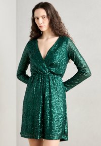 Abito verde in paillettes con scollatura a V profonda, maniche lunghe e vita affusolata, caratterizzato da una superficie testurizzata e riflettente e orlo svasato.