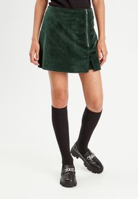 Green corduroy skirt with a front zipper, featuring a high slit. Paired with black knee-high socks and chunky black loafers with a chain detail.