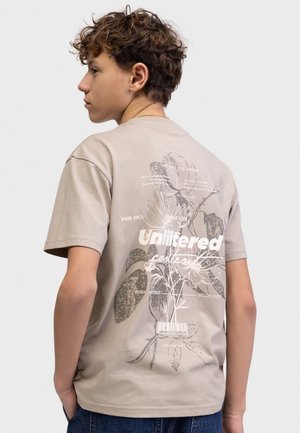 Έφηβος με σγουρά μαλλιά φοράει μπεζ μπλουζάκι με μεγάλο λουλουδάτο σχέδιο και το κείμενο "Unfiltered" στην πλάτη, με τα χέρια στις τσέπες, κοιτάζοντας προς τα αριστερά.