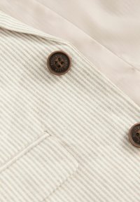 Gros plan sur un tissu beige et blanc rayé avec deux boutons marron étiquetés "AUTHENTIC NEXT DUDE" et une petite poche.