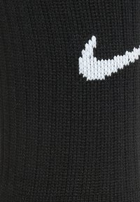 Sort strikket sok med en tekstureret overflade, der har et hvidt Nike swoosh-logo. Stoffet ser holdbart og strækbart ud.