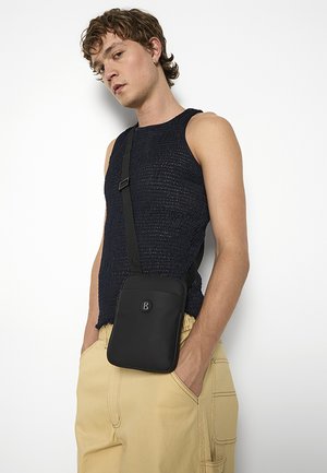 Borsa a tracolla nera realizzata in materiale liscio con logo, caratterizzata da una tracolla regolabile e un design minimalista; indossata sopra una canotta blu navy con texture.