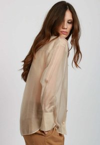 Camicia button-up beige traslucida con una vestibilità rilassata, maniche lunghe e orlo curvo. La sua texture morbida e la leggera sottigliezza ne esaltano l'aspetto.