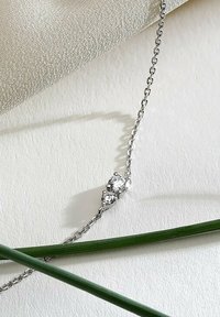 Silver chainhalsband med två klara runda ädelstenar, placerade i en enkel, elegant design, som vilar på en texturerad tygbakgrund.