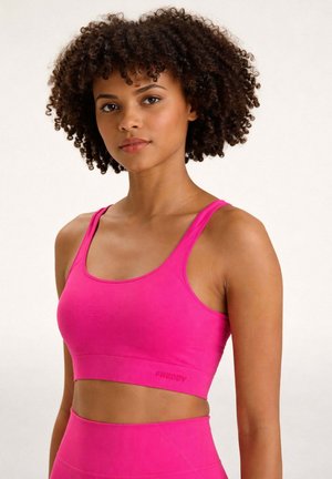 Junge Frau mit lockigem Haar, die einen leuchtend pinken Sport-BH und dazu passende, hoch taillierte Leggings trägt, stehend vor einem schlichten, hellen Hintergrund.