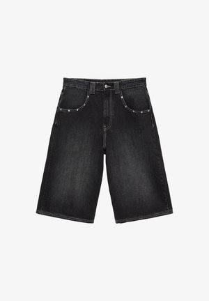 Ung mand iført en løs hvid T-shirt, oversized sorte denimshorts med store lommer, hvide sokker og sorte sneakers, stående mod en almindelig væg.