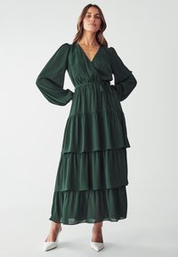 Green, tiered maxi dress made of soft fabric. Features a v-neckline, long sleeves, and a tie waist detail. Styled with white high heels.