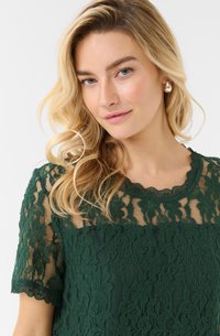 Top in pizzo verde con maniche corte, scollatura a onde e motivo floreale con sovrapposizione trasparente, evidenziando dettagli e trame intricati.