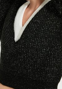 Crop top noir en maille avec une finition texturée, doté d'un col en V blanc. Le tissu présente un léger éclat et un motif de tissage complexe.